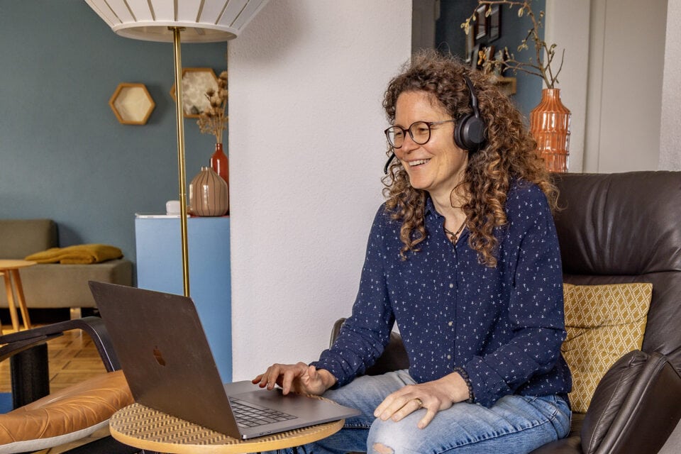 Frau mit Laptop und Kopfhörer, lächelnd im Wohnzimmer.